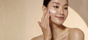 A close-up of a woman with dark hair, wearing a sheer nude strapless top, applying a white cream to her cheek against a warm beige background.
