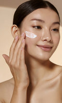 A close-up of a woman with dark hair pulled back, applying a white cream product to her cheek with her finger, set against a soft beige background.