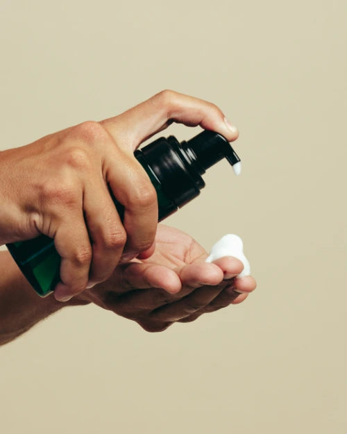 Hand holding a skincare bottle with a pump dispensing cleanser to another hand against a beige background