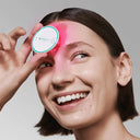 Woman holding a pink omnilux led face device spot blemish device over her face with a gray background