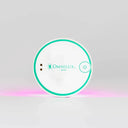 A round white Omnilux Mini LED device with a teal ring border and logo, resting on a reflective surface with a pink glow underneath, set against a light gray background.