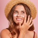 Woman wearing a straw hat with sunscreen on her face against a pink background