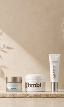 Three skincare products from the brands hmbl, Alastin, and Skinceuticals are lined up on a textured beige stone surface against a matching beige plaster wall with soft leaf shadows.