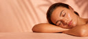 A woman with closed eyes rests her head on her arm on a table, bathed in warm peach and pink lighting against a softly blurred spa-like background.