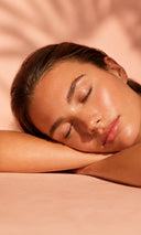 A close-up lifestyle shot of a woman with closed eyes resting her head on her arms, showcasing a glowing, hydrated complexion against a warm peach background with soft leaf shadows.