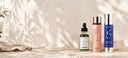 Three skincare products from Skinceuticals, skinbetter science and ZO Skin Health are lined up on a textured beige stone surface with soft leaf shadows cast across them. The products include a green dropper bottle, a pink bottle with a gold cap, and a blue-and-silver bottle.