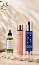 Three skincare products from SkinCeuticals, skinbetter science and ZO Skin are arranged on a light stone tray against a textured beige wall with soft leaf shadows. The products include a green dropper bottle, a peach-pink pump bottle with a rose gold cap, and a blue cylindrical bottle with a silver cap.